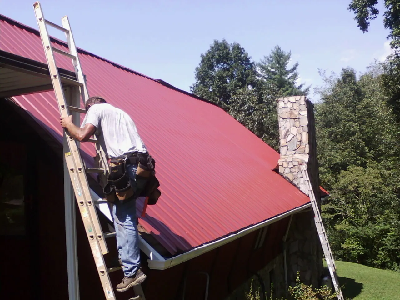 Roofer on a ladder beside a red metal roof and stone chimney — Soffit & Fascia Repair by Roofing Brothers in Zarephath